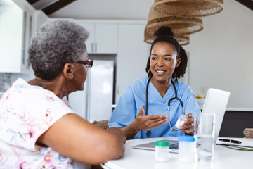 Smiling african american doctor with laptop on table giving medicines to senior patient at home