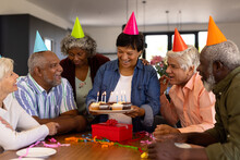 Multiracial Seniors Wearing Hats Looking At Happy Woman Holding Cupcakes With Birthday Candles