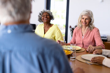 Wall Mural - Happy multiracial senior friends talking while sitting at dining table in nursing home