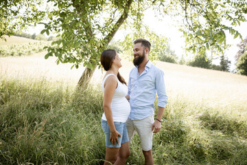 Wall Mural - pretty young pregnant woman with white shirt stands with her boyfriend with beard and blue shirt in high flower meadow and cuddle