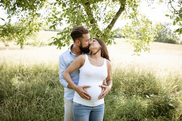 Wall Mural - pretty young pregnant woman with white shirt stands with her boyfriend with beard and blue shirt in high flower meadow and cuddle