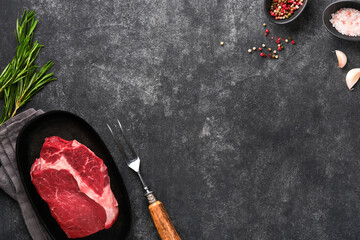 Wall Mural - Beef steak. Fresh raw beef rib eye steak with fork rosemary, salt and pepper on a grill pan on dark grey or black stone background. Shop advertising idea. Top view. Mockup for design idea.