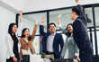 Group of Asian people wearing formal business suits standing together, raising hands with happiness, celebrating on success in meeting room of indoor office or workplace.