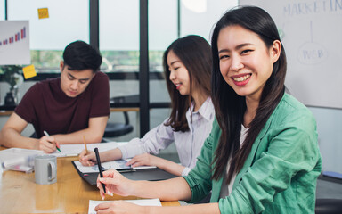 Selective focus foreground of adorable casual business woman smiling with happiness and confidence while sitting in meeting room with Asian colleagues for sharing creative ideas in modern office.