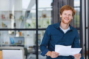 Smiling successful businessman stands near desk smiling and looking at the camera. Young positive male confident entrepreneur or small business owner in a modern office or coworking space