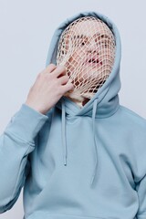 Wall Mural - a man with a mesh on his face stands in a light blue hoodie on a light background and tries to remove it from his face