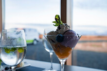 Close-up Of Tempting Dessert Served In Glass. Tasty Ice Cream And Fruit Mix With Wineglass Arranged On Dining Table. View Of Sky Against Window At Restaurant In Hotel.
