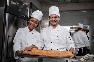 Wall Mural - Portrait of professional chefs in white uniform looking at camera with a cheerful smile and proud with tray of bread in kitchen. A friend and partner of pastry foods and fresh daily bakery occupation.