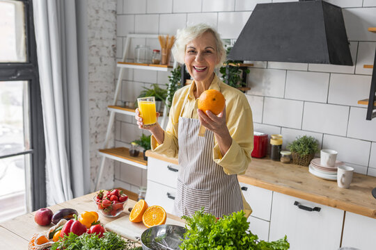 Attractive senior woman drinking freshly squeezed orange juice, looking happy and healthy. Concept of active lifestyle of mature people, cooking organic food. Mature female at home at her kitchen