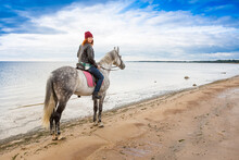 Wearing Jeans, Jacket And Warm Hat Woman Rides Astraddle Grey Horse Along Sandy Beach