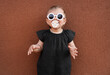 Cute little girl wearing stylish clothes with sunglasses and pacifier near brown wall