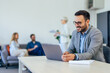 © bnenin - Adult businessman working over the laptop while his colleagues talking in the background.