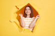 © sementsova321 - Horizontal shot of smiling happy woman wearing striped shirt posing in yellow paper hole, standing with raised arms, celebrating her success, looking at camera with toothy smile.