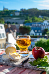 Products of Normandy, cow neufchatel lait cru cheese and glass of apple cider drink with houses of Etretat village on background, Normandy, France