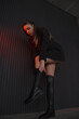 © NASTYA PALEHINA - teen leisure. young girl in black slim dress-jacket is standing fashion and pulling over the knee boots near on loft wall background with red studio light. bottom camera. lifestyle concept, free space