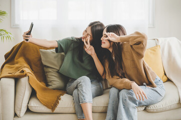 Wall Mural - Two asian young women happy smiling and taking selfie on couch in living room at home