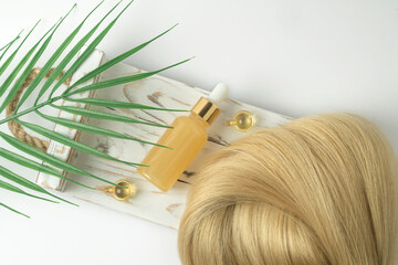 Natural blonde hair strand and essential oil for hair treatment in golden capsules lying on a white wooden tray