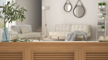 Wooden table top, cabinet, panel or shelf with shutters close up. Olive branch in vase and candles. Blurred background with white living room with sofa and armchairs, interior design