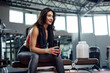 A fit, muscular sportswoman is sitting on a bench in a gym and drinking protein shake.