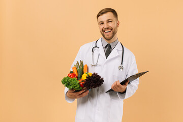 The happy male doctor nutritionist with stethoscope holding fresh vegetables on beige background, diet plan concept