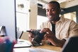 © Southworks - Young Black Professional Photographer Sitting At Desk Working On Computer Holding Camera Editing Pictures