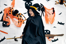 Halloween Wizard And Witch Concept - Senior Woman In Witch Costumes Celebrating Halloween Laughing On Broom With Bats And Spider Web Over White Background