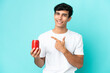 © luismolinero - Young Argentinian man holding a refreshment isolated on blue background pointing to the side to present a product
