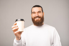 Portrait Of A Smiling Bearded Man Holding And Giving You A Cup Of Coffee. Sudio Shot Over Grey Background.