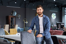 Successful Store Owner In Casual Clothes Leaning On Table, While Posing In Furniture Store. Portrait Of Confident Businessman Looking At Camera And Smiling In Exhibition Center. Concept Of Business.