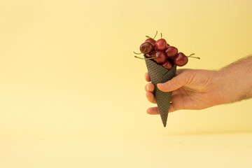 Wall Mural - Trendy summer composition with fresh red cherries and a black ice cream cone in a man's hand against a pastel yellow background. Minimal summer food idea.