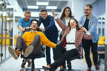 Young Cheerful Business People Racing On Office Chairs And Laughing. Funny Activity, Celebrating Corporate Success. Business Concept.