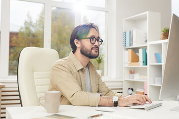 Content creative young Asian man with beard sitting at desk and typing on keyboards while communicating with client online