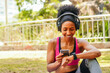 Attractive young black woman exercising on sports mat in city park, exercising her muscles watching her smarwatch and using her smartphone, leading active lifestyle outdoors