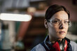 © Mediaphotos - Portrait of serious attractive tough woman with brown hair wearing red ear protectors on neck standing at factory workshop