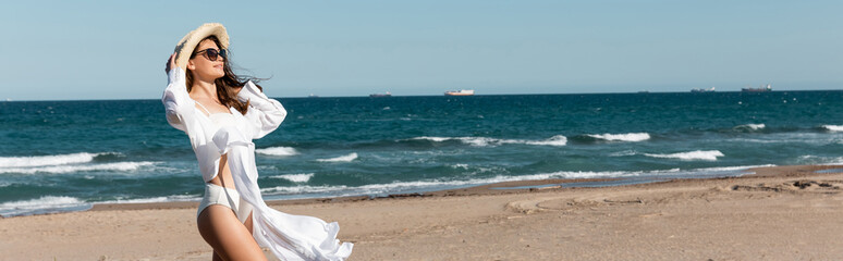 Canvas Print - smiling woman in sunglasses and straw hat standing in white shirt near sea on beach, banner.