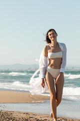 Wall Mural - happy young woman in white shirt and swimwear walking on sea shore.