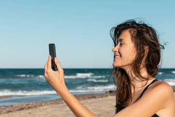 Wall Mural - cheerful young woman in swimsuit taking selfie on smartphone near sea.