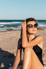 Canvas Print - sunshine on face of young woman in black swimwear and sunglasses sitting on beach near sea.