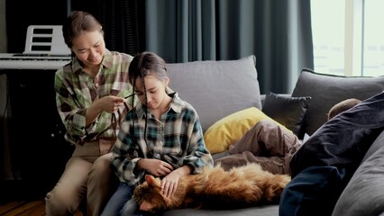 Mom braids her daughter's hair in the morning, sitting on the sofa in the living room. Girl petting a big red Maine Coon cat