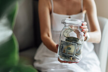 Young Caucasian Woman Holding Big Glass Bottle With Cash Money Dollar Bill On White Marble Table At Home. Investment Business, Retirement, Finance And Saving Money For Future Concept.
