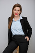 Smiling woman in black business suit sitting on tall stool and holding one hand in pants pocket. Young business woman isolated portrait.