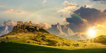 Castle On The Hill Beneath A Rainbow At Sunset. Composite Fantasy Landscape. Grassy Meadow In The Foreground. Rocky Peaks Of The Ridge In The Distant Background In Evening Light