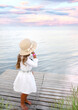 © Alina - A little Caucasian girl in a white dress and hat, view from the back, listens to the sea. Background of a pink-gray sunset. Relaxation. Childhood concept