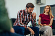 © bnenin - Close-up of a depressed young man sharing problems sitting in a circle during a group therapy session.