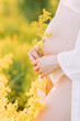 Pregnant woman stands in unbuttoned white shirt and bikini among yellow flowers.