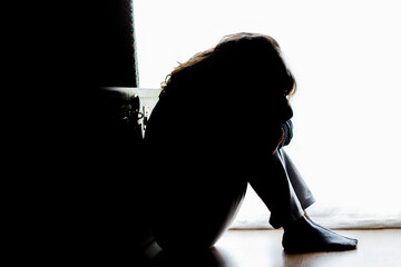 Unhappy mature lady, woman in silhouette, sitting on the floor with back lit. Depressing thoughts. 