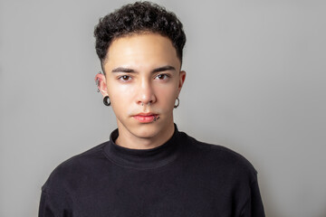 Attractive young man with curly hair, wearing piercings, looking at camera over gray background with copy space.