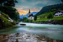 The Parish Church Of St. Sebastian In Ramsau Near Berchtesgaden As A Long Exposure With The River And A Bridge At Sunset