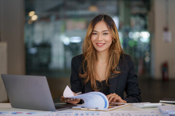 Wall Mural - Smiling beautiful young Asian businesswoman sitting with laptop and computer while doing some paperwork at the office. Looking at camera.