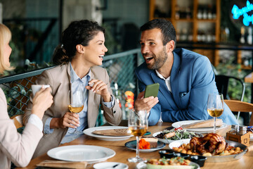 Cheerful colleagues have fun while using cell phone during business lunch in restaurant.